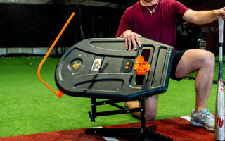A man in a red shirt and black shorts with an aluminum bat kneels behind the swing path trainer in a batting cage.