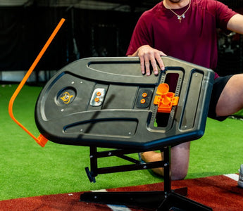 A man in a red shirt and black shorts with an aluminum bat kneels behind the swing path trainer in a batting cage.