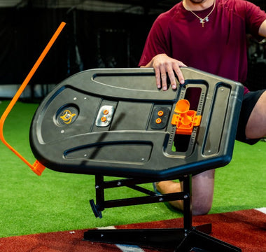A man in a red shirt and black shorts with an aluminum bat kneels behind the swing path trainer in a batting cage.