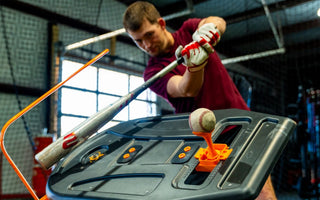 A close-up of a man in a red shirt swinging a baseball bat through a swing trainer at a ball on a tee in a batting cage.