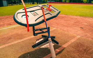 A close-up of the Perfect Swings USA Lag Bat leaning against the Swing Path Trainer over home plate of a baseball field.