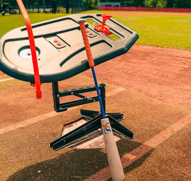 A close-up of the Perfect Swings USA Lag Bat leaning against the Swing Path Trainer over home plate of a baseball field.