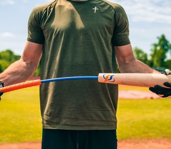 A man in a green T-shirt holding the Perfect Swings USA Lag bat by both ends and bending it on a baseball diamond.
