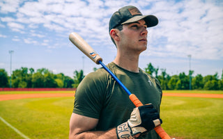 A baseball player holds a bat on a sunny field, looking into the distance before practice or a game begins.