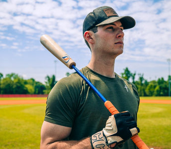 A baseball player holds a bat on a sunny field, looking into the distance before practice or a game begins.