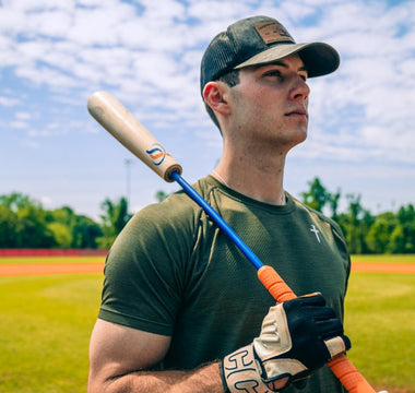 A baseball player holds a bat on a sunny field, looking into the distance before practice or a game begins.