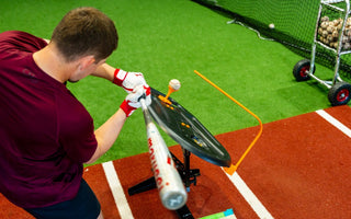 A man in a maroon shirt swinging a baseball bat toward a ball on a tee inside an indoor training area.