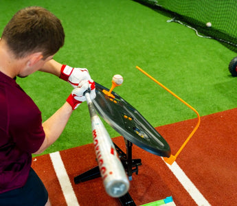 A man in a maroon shirt swinging a baseball bat toward a ball on a tee inside an indoor training area.