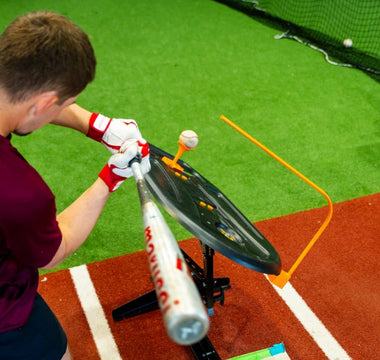 A man in a maroon shirt swinging a baseball bat toward a ball on a tee inside an indoor training area.